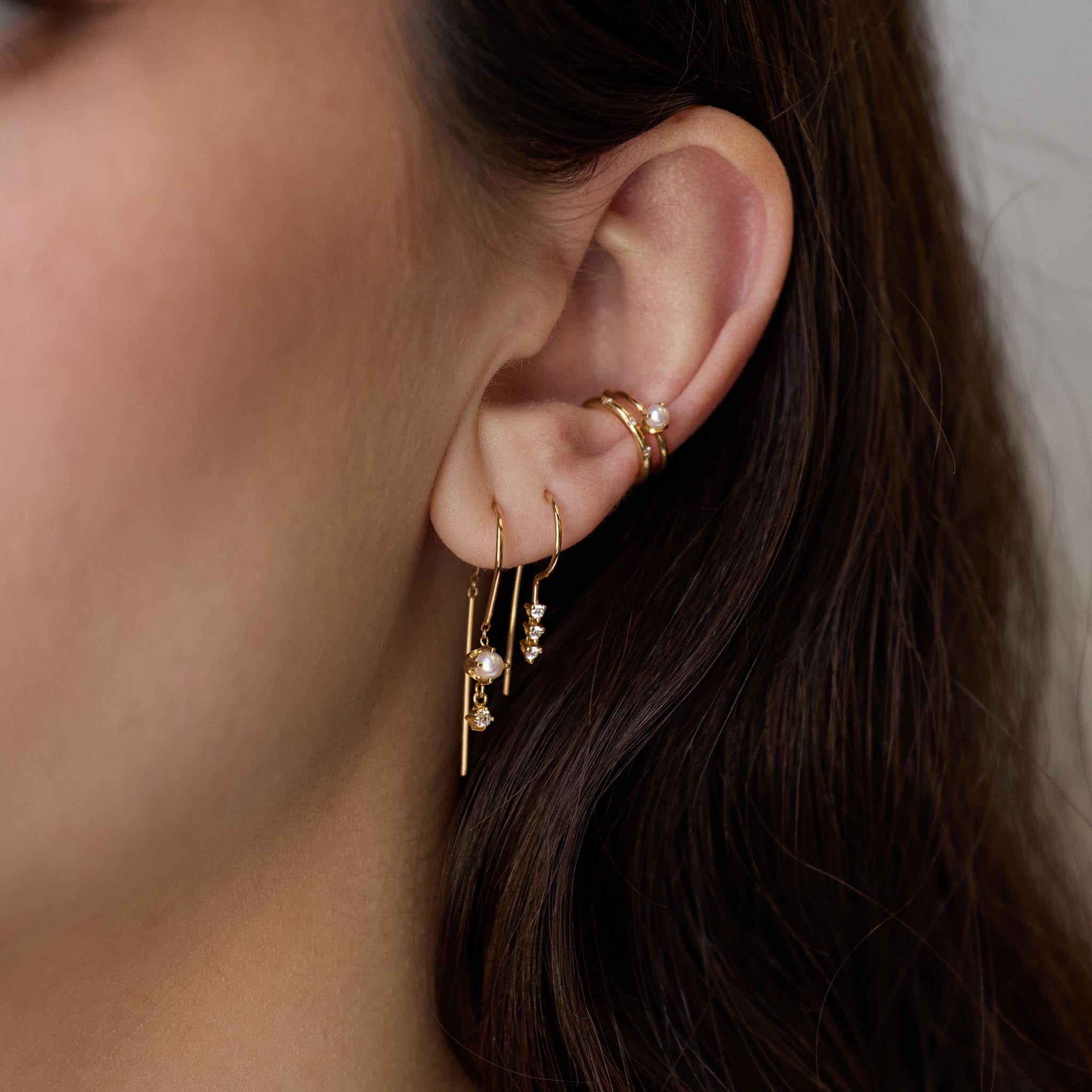 Close up of a brunette woman's ear wearing a Zoë Chicco 14k Gold Dangling Pearl & Diamond U Wire Threader Earring layered with a 14k Diamond Tennis Wire Hook Earring in her second piercing, a Pearl Ear Cuff, and a 14k Spread Out Bead Set Diamond Thick Ear Cuff
