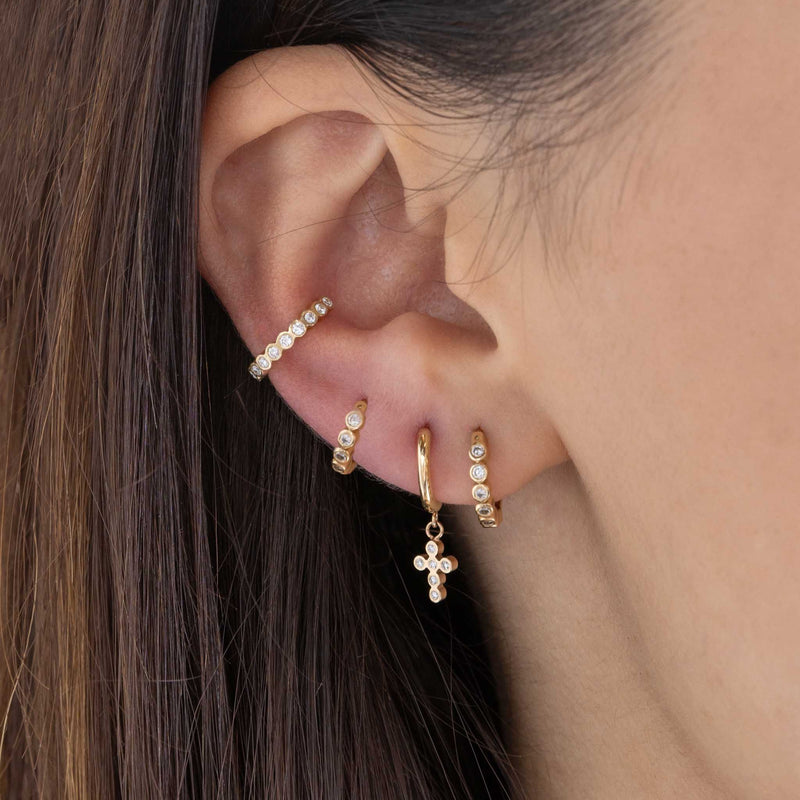 close up of a woman's ear wearing two sizes of Zoë Chicco 14k Gold Diamond Bezel Small Hinge Huggie Hoop Earrings in different piercings layered with a 14k Dangling Diamond Bezel Cross Small Hinge Huggie Hoop and a Diamond Bezel Ear Cuff