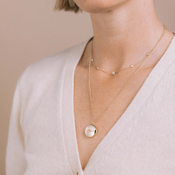 woman in a white cardigan sweater wearing a Zoë Chicco 14k Gold Floating Graduated Diamond Station Necklace layered with a 14k Gold Grateful Round Locket Necklace