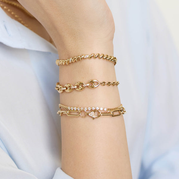 Close up of a wrist wearing multiple Zoe Chicco 14k gold chain and diamond bracelets on a light blue background