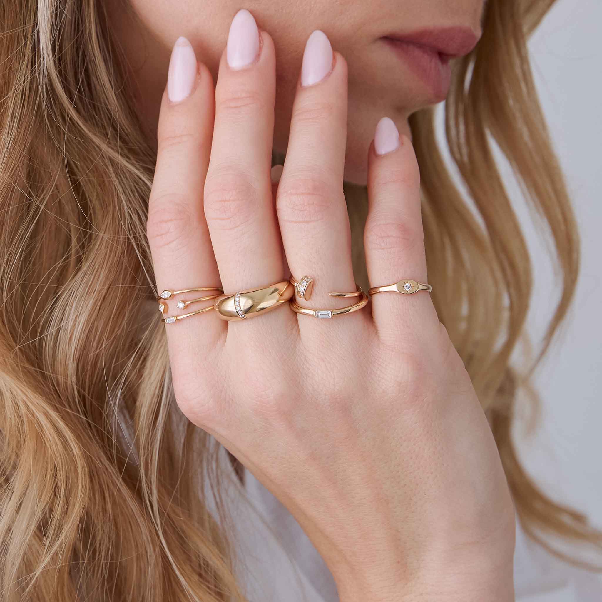 woman with her hand raised to her cheek wearing a Zoë Chicco 14k Gold Pavé Diamond Nail Open Ring stacked with a 14k Baguette Diamond Thick Band Ring on her ring finger, a 14k Star Set Diamond Oval Signet Ring on her pinky, a 14k 3 Thin Band Mixed Diamonds Ring on her index, and a 14k Pavé Diamond Line Small Aura Ring on her middle finger