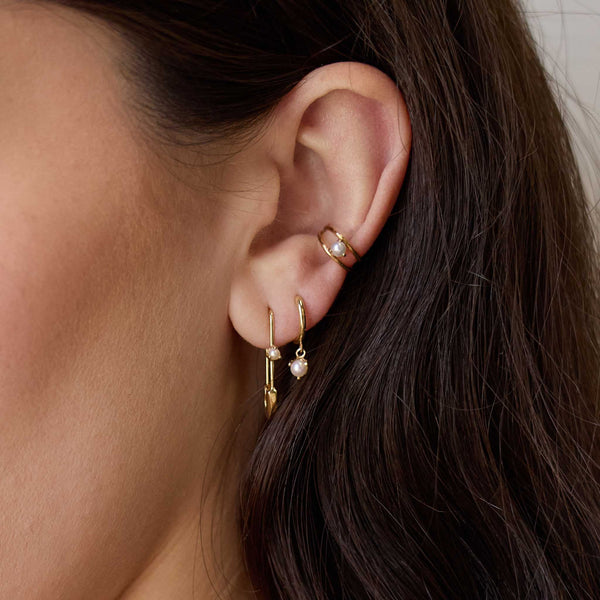 close up of a woman's ear wearing a Zoë Chicco 14k Gold Dangling Pearl Small Hinge Huggie Hoop Earring layered with a 14k Gold Safety Pin with Pearl Threader Earring and a 14k Pearl Double Wire Ear Cuff
