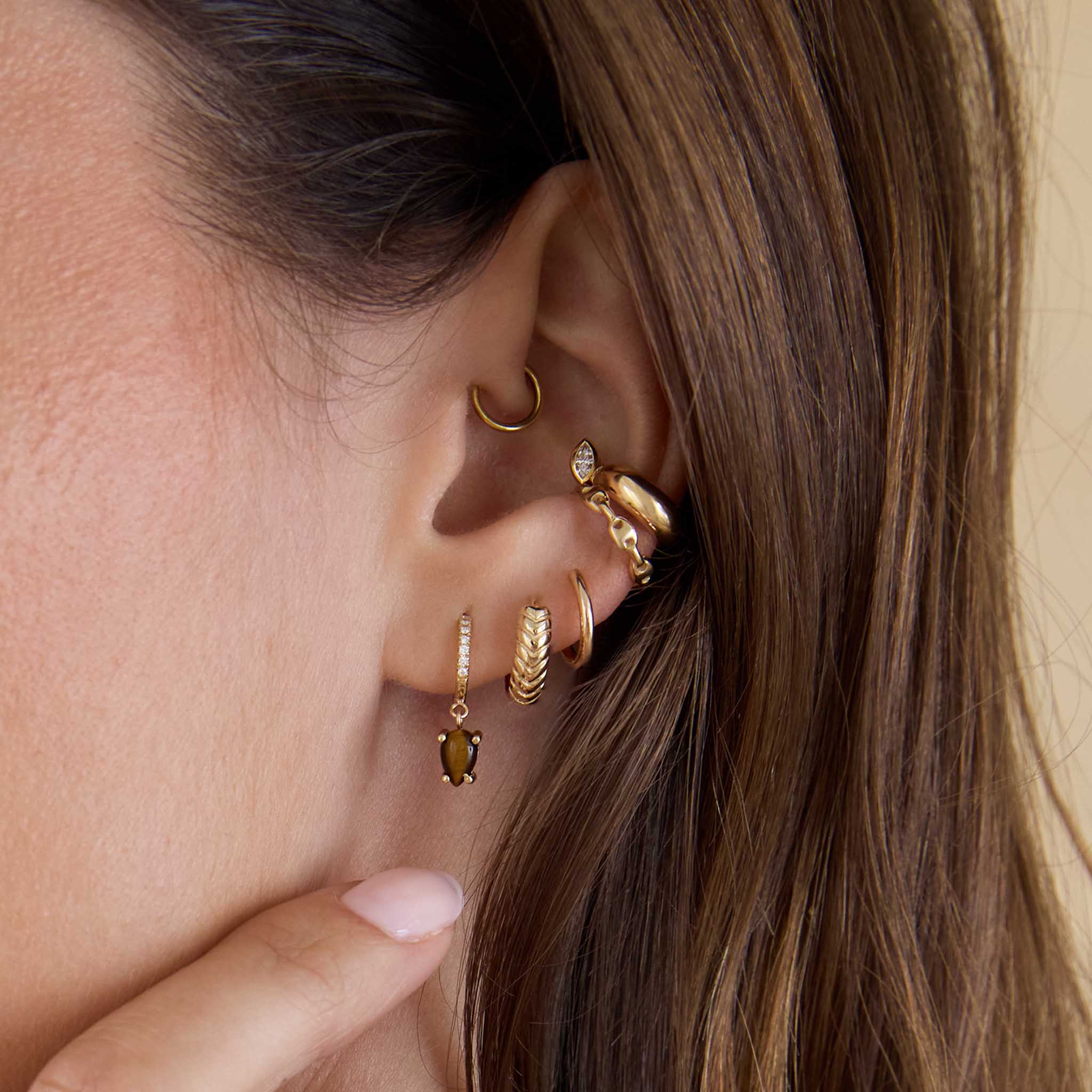 Close up of a woman's ear wearing a Zoë Chicco 14k Gold Small Mariner Chain Ear Cuff and Marquise Diamond Chubby Ear Cuff layered with a 14k Dangling Pear Tiger Eye Small Pavé Diamond Hinge Huggie Hoop and Tapered Braided Huggie Hoop
