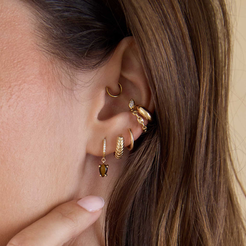 Close up of a woman's ear wearing a Zoë Chicco 14k Gold Small Mariner Chain Ear Cuff and Marquise Diamond Chubby Ear Cuff layered with a 14k Dangling Pear Tiger Eye Small Pavé Diamond Hinge Huggie Hoop and Tapered Braided Huggie Hoop