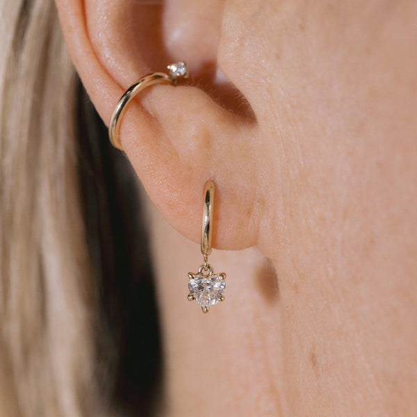 close up of a woman's ear wearing a Zoë Chicco 14k Gold Dangling Heart Diamond Small Hinge Huggie Hoop Earring