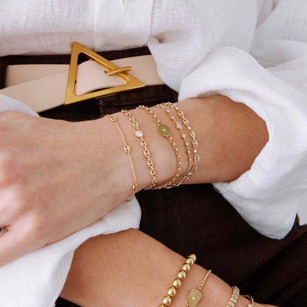 woman in a white shirt and brown pants  with her arms in her lap wearing a Zoë Chicco 14k Gold Small Mariner Link Station Bracelet stacked with a 14k Floating Diamond Small Puffed Mariner Chain Bracelet, 14k Star Set Diamond Disc Rolo Chain Bracelet, 14k Marquise & Graduated Round Diamond Bolo Bracelet, and a 14k 5 Prong Diamond Small Puffed Mariner Chain Bracelet