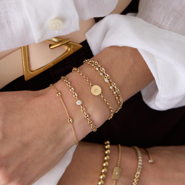 woman in a white shirt and brown pants  with her arms in her lap wearing a Zoë Chicco 14k Gold Small Mariner Link Station Bracelet stacked with a 14k Floating Diamond Small Puffed Mariner Chain Bracelet, 14k Star Set Diamond Disc Rolo Chain Bracelet, 14k Marquise & Graduated Round Diamond Bolo Bracelet, and a 14k 5 Prong Diamond Small Puffed Mariner Chain Bracelet