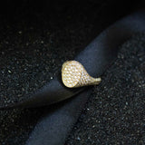 A pave diamond encrusted signet ring is sitting on black ribbon and black sand.  The ring has an oval face and is often styled as a pinky ring.