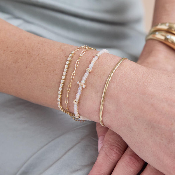 a woman's wrist resting on her lap wearing a Zoë Chicco 14k Gold Small Snake Chain Bracelet stacked with a 14k Gold & Fire Opal Rondelle Bead Bracelet with 2 Prong Diamonds, 14k Gold Small Paperclip Chain Bracelet and a 2mm Diamond Bezel Tennis Bracelet