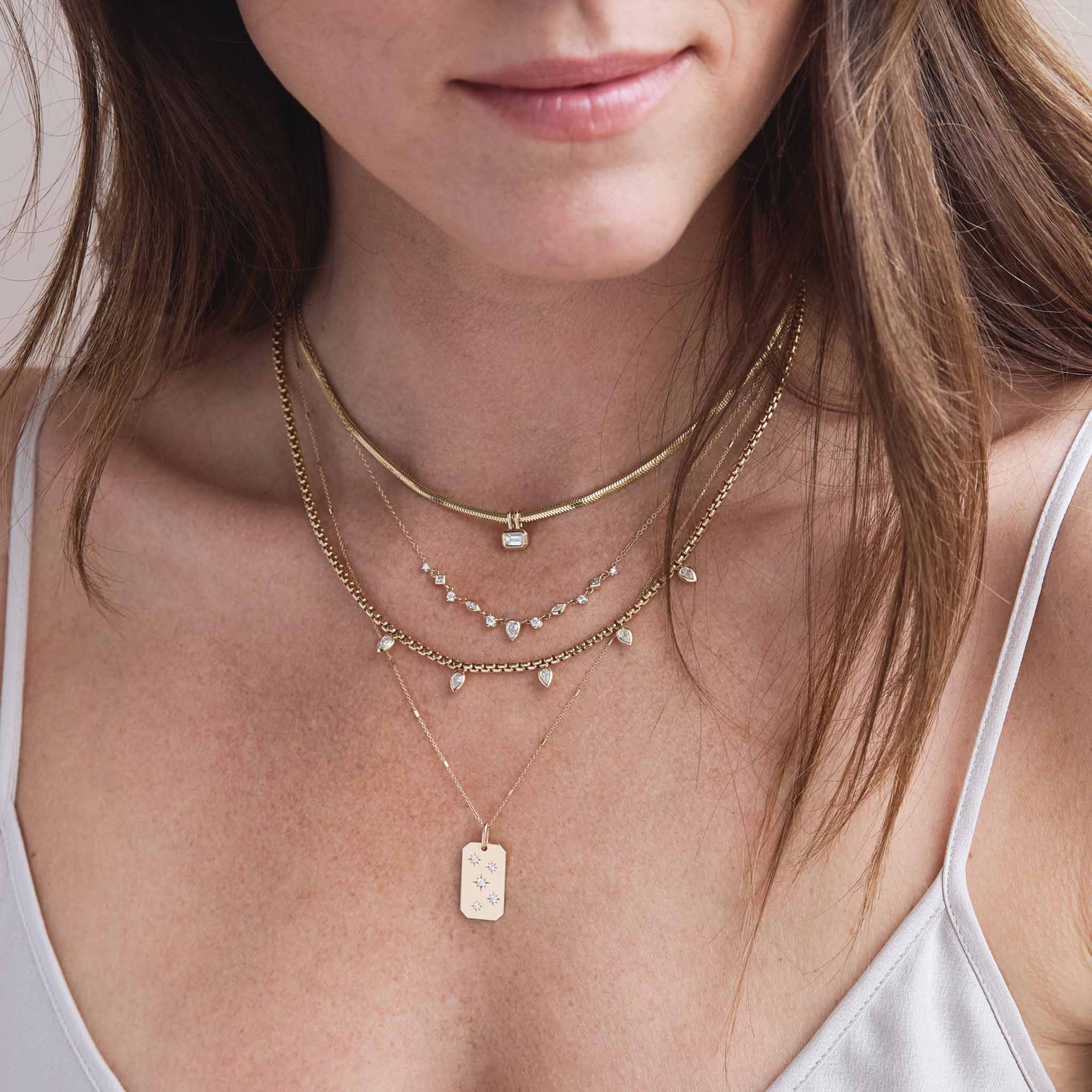 woman in a gray camisole top wearing a Zoë Chicco 14k Gold 11 Linked Mixed Fancy Cut Diamond Necklace layered with a 14k Emerald Cut Diamond Pendant Snake Chain Necklace, 14k 5 Vertical Pear Diamond Medium Box Chain Necklace, and a 14k Scattered Star Set Diamonds Square Edge Dog Tag Necklace