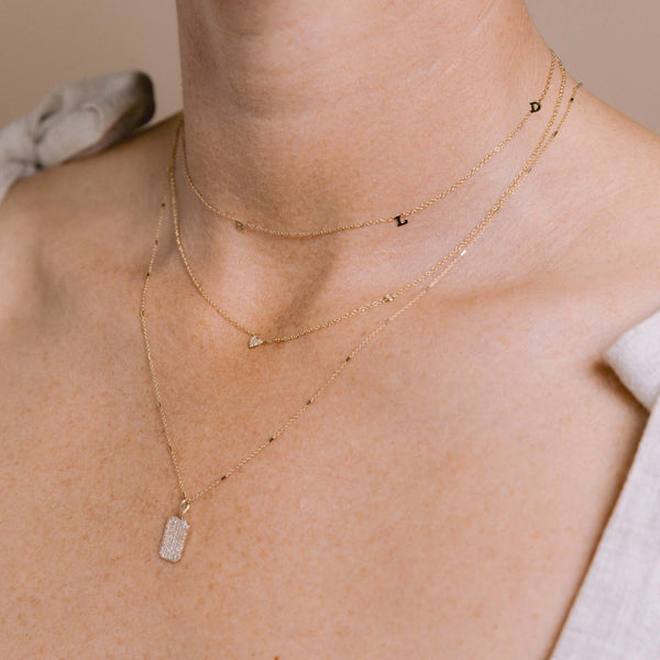 woman wearing a Zoë Chicco 14k Gold Itty Bitty Multi Letter Station Necklace customized with four initials layered with a 14k Itty Bitty Pavé Diamond Heart Necklace with Floating Diamond and a 14k Pavé Diamond Small Square Edge Dog Tag Necklace