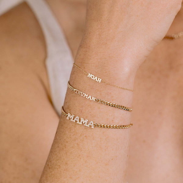 woman's wrist wearing a Zoë Chicco 14k Gold Custom Itty Bitty Letters Bracelet with the name NOAH and a Zoë Chicco 14k Gold Custom Itty Bitty Letters Curb Chain Bracelet with the name TRUMAN layered with a Pave Diamond MAMA Small Curb Chain Bracelet