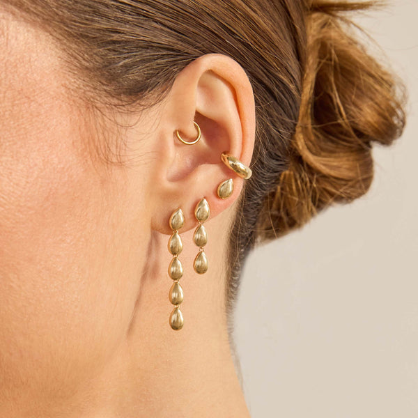 Close-up of a person wearing Zoe Chicco 14k gold hoop earrings and teardrop shaped dangling earrings with a neutral background
