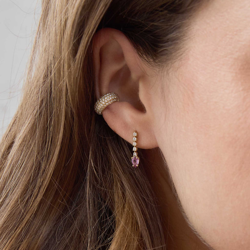 Close-up of an ear wearing Zoe Chicco 14k gold pave ear cuff and a single dangle earring with diamonds and a pink pear-shaped sapphire on a neutral background