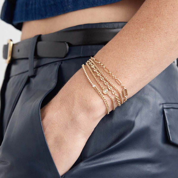 A woman with navy leather pants has her hand in her pocket and is wearing stack of Zoe Chicco Bracelets including: 14k Gold Medium Box Chain Bracelet, 14k Gold Small Puffed Mariner Chain Bracelet, 14k Diamond Nugget Box Chain Bracelet and 14k Floating Diamond Mixed Curb Chain & Diamond Tennis Bracelet