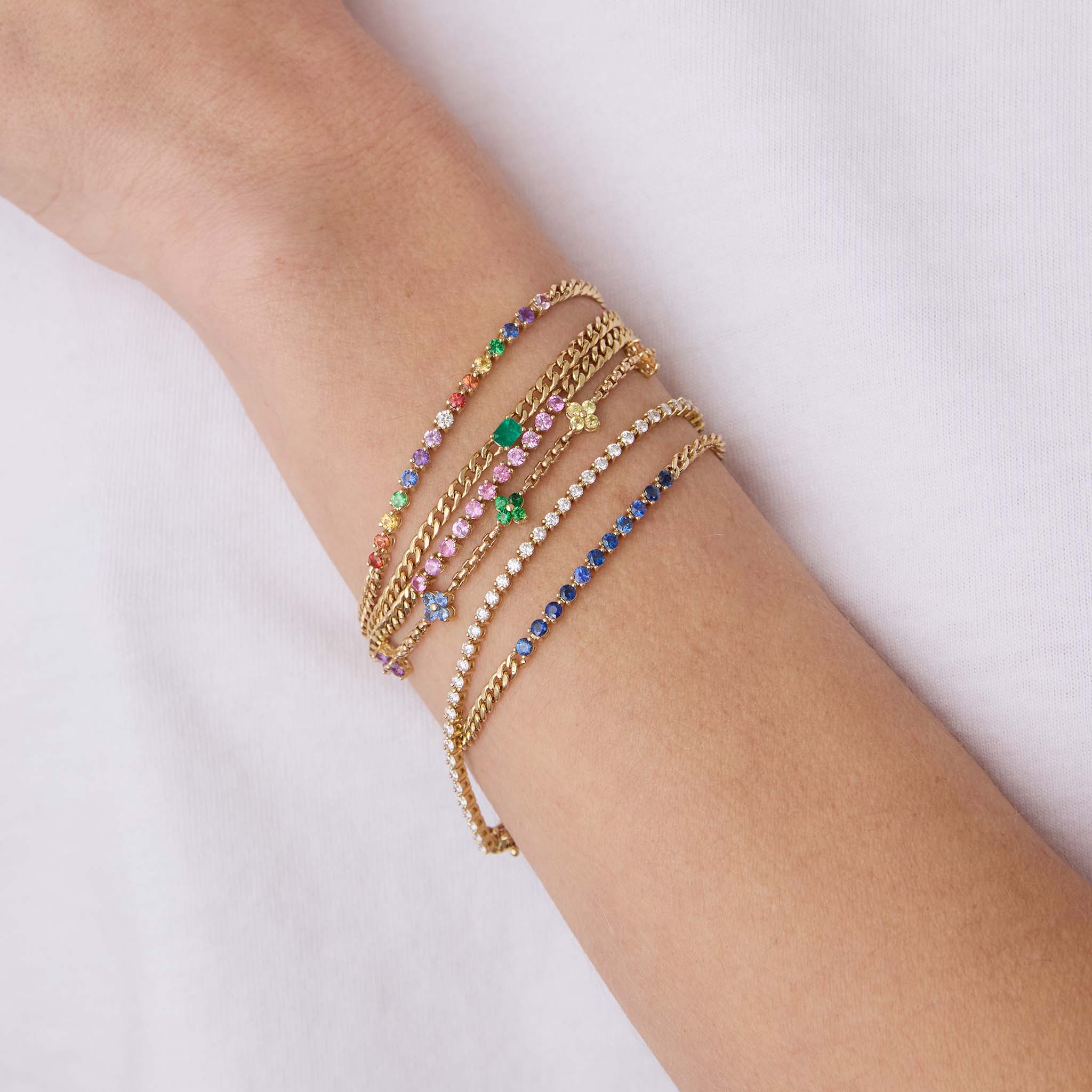 woman's wrist wearing a Zoë Chicco 14k Gold Rainbow Sapphire Tennis Segment Small Curb Chain Bracelet stacked with a 14k Emerald Cut Emerald Small Curb Chain Bracelet, Pink Sapphire and Blue Sapphire Tennis Small Curb Chain Bracelets, a Prong Diamond Tennis Bracelet, and a 14k Rainbow Sapphire Quad Station Box Chain Bracelet