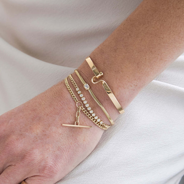 close up of a woman's hand resting in her lap wearing a Zoë Chicco 14k Gold Emerald Cut Diamond Small Snake Chain Bracelet layered with a Diamond Bezel Tennis Segment Curb Chain Bracelet and a Small Curb Chain Bar Toggle Bracelet