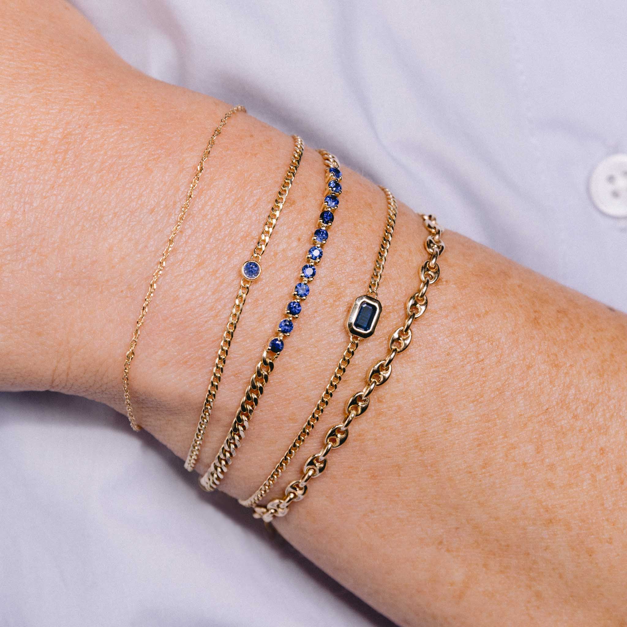 woman's wrist wearing a Zoë Chicco 14k Gold Blue Sapphire Bezel XS Curb Chain Bracelet stacked with a 14k Emerald Cut Blue Sapphire XS Curb Chain Bracelet, 14k Blue Sapphire Tennis Segment Small Curb Chain Bracelet, and a Small Mariner Chain Bracelet