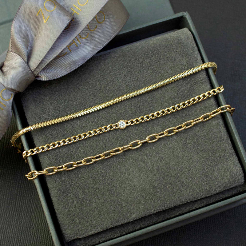 Inside a grey jewelry box are 3 bracelets including the small snake chain, small curb with floating diamond and the medium square oval link bracelets in 14k yellow gold.