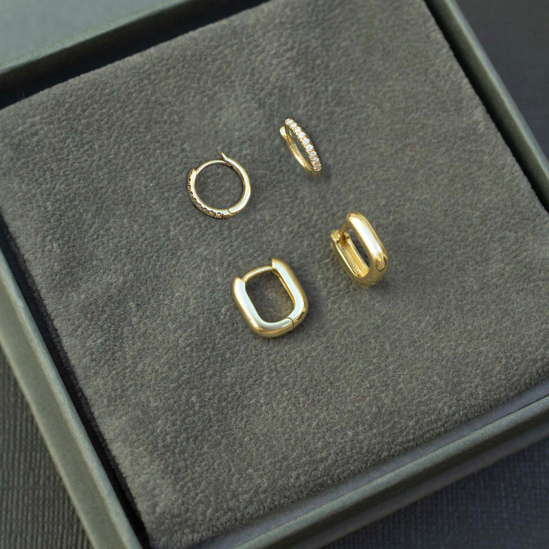 On top of a gray box is two pairs of huggie hoop earrings:  small pave diamond huggies and the small thick oval huggies.