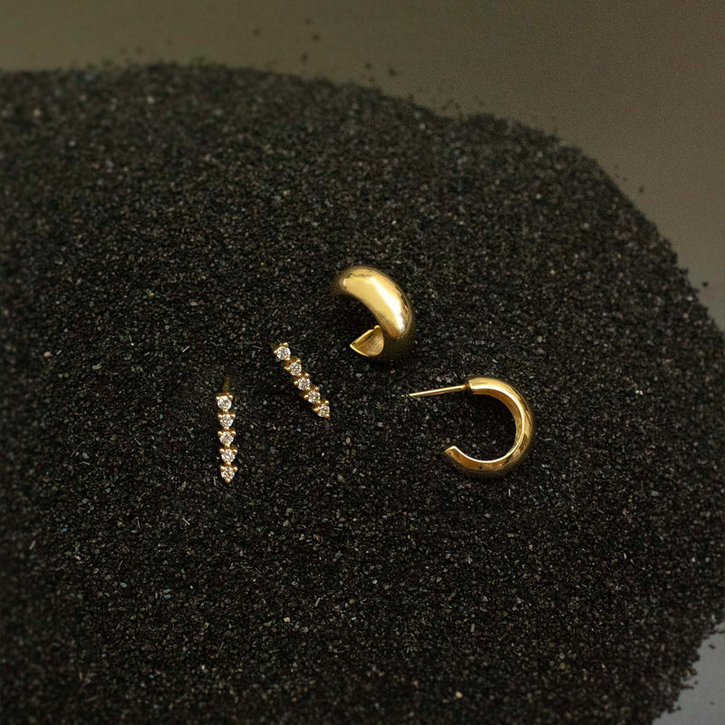 Resting on black sand are two pairs of earrings:  diamond tennis drops and chubby huggies.  Both earrings are 14k yellow gold.