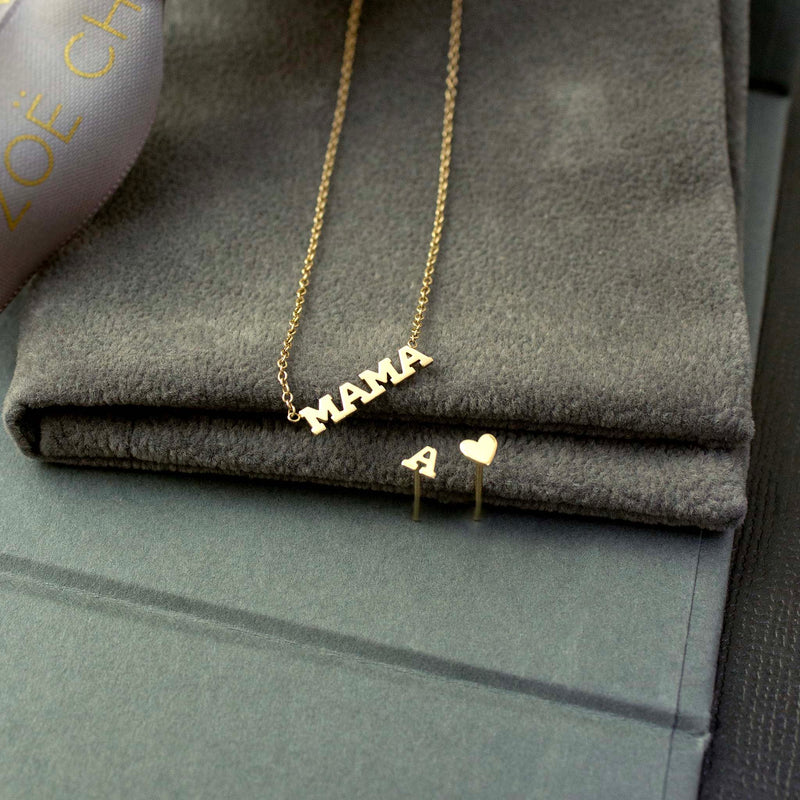 Laying on a gray background is our best selling MAMA necklace and a single itty bitty heart stud and tiny letter stud.