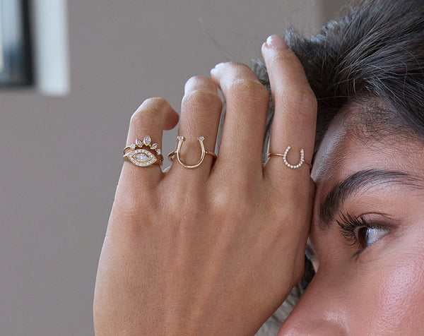 Close-up of a hand wearing Zoe Chicco 14k gold and diamond horseshoe rings against a blurred grey background