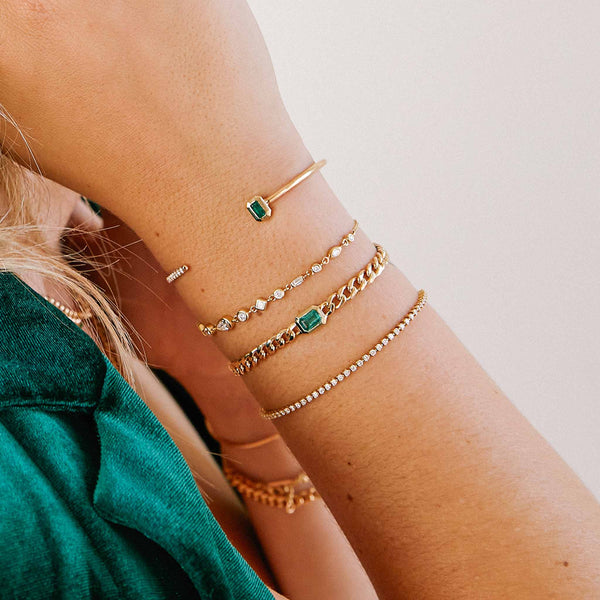 cropped view of a wrist wearing a Zoë Chicco 14k Gold Medium Curb Chain Emerald Cut Emerald Bezel Bracelet layered with three other bracelets