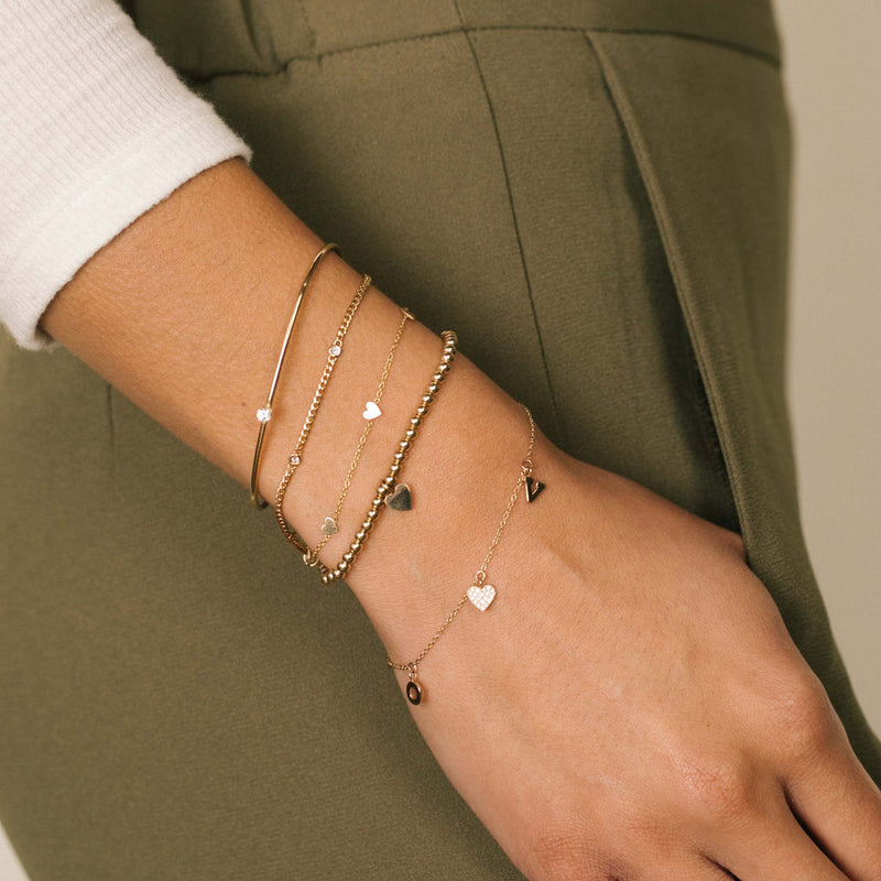 woman in white top and olive pants wearing Zoë Chicco 14k Gold Single Prong Diamond Cuff Bracelet on her wrist layered with 5 Itty Bitty Heart Bracelets and Small Gold Bead Midi Bitty Heart Bracelet