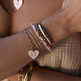 close up of woman's wrist wearing Zoë Chicco 14k Gold & Faceted Pink Opal Rondelle Bead Bracelet with 2 Diamonds layered with a Graduated Prong Diamond Tennis Bolo Bracelet, Diamond Wide Half Round Cuff, LOVE Square oval Link Bracelet, and a Princess Diamond and Curb Chain Double Bracelet