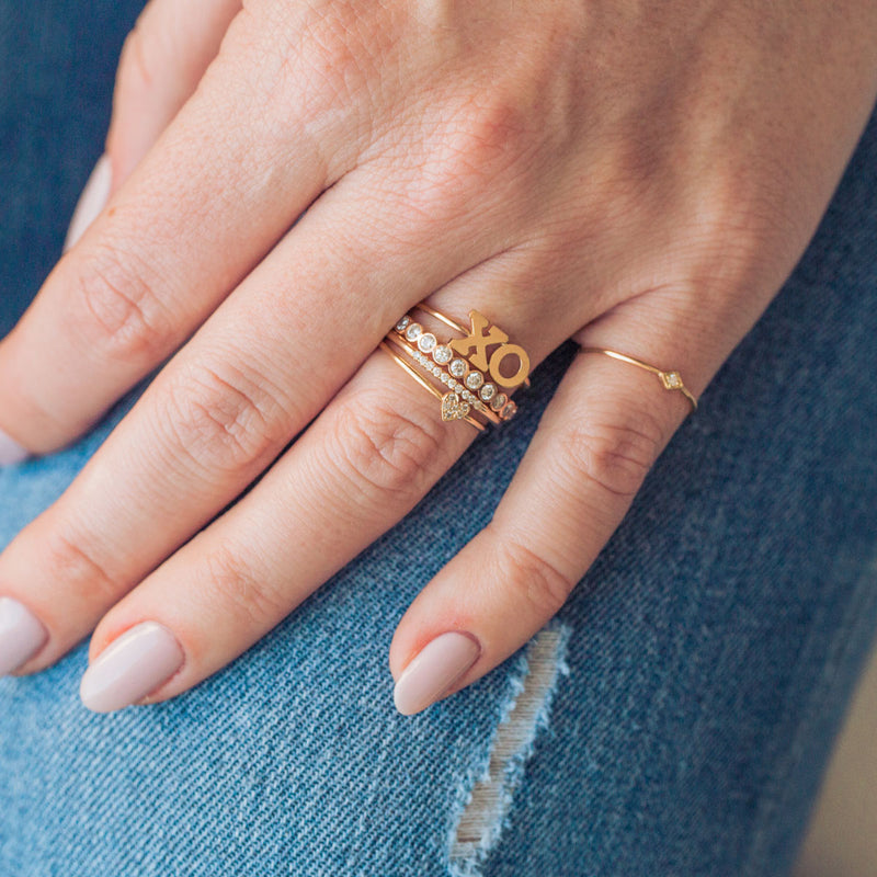 woman's hand resting on jeans wearing an XO letter ring, Diamond Bezel Eternity Band, 10 Pave Diamond Medium Band, and an Itty Bitty Pave Diamond Heart Ring stacked together