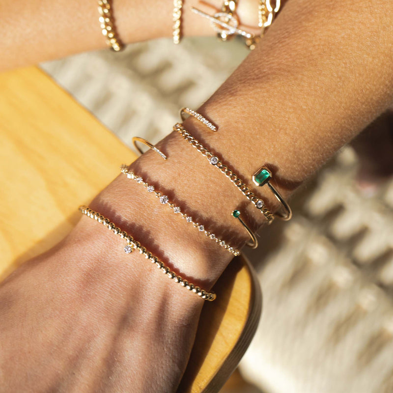 close up of wrist resting on table wearing Zoë Chicco 14kt Small Gold Bead with Diamond Bezel Charm Bracelet layered with two emerald and diamond cuffs and a Graduated Prong Diamond Bolo Bracelet and Floating Diamond Curb Chain Bracelet