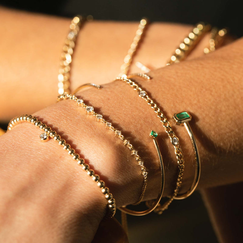 close up of wrist wearing Zoë Chicco 14kt Small Gold Bead with Diamond Bezel Charm Bracelet layered with two emerald and diamond cuffs and a Graduated Prong Diamond Bolo Bracelet and Floating Diamond Curb Chain Bracelet