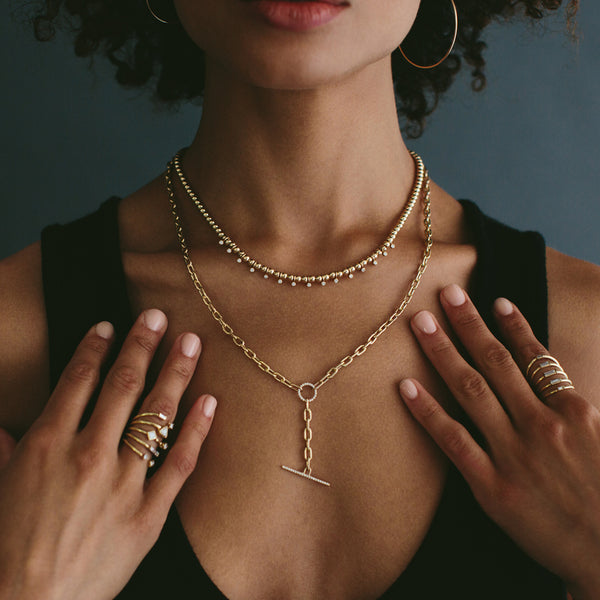 woman standing against a dark background wearing a Zoë Chicco 14k Gold Bead Eternity Dangling Diamond Bezel Necklace layered with another necklace