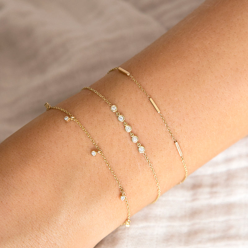 woman's wrist wearing Zoë Chicco 14kt Gold 5 Dangling Diamond Bezel Bracelet layered with a linked floating diamond bracelet and a tiny bar bracelet