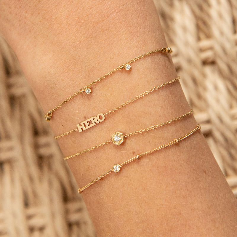 woman's wrist wearing Zoë Chicco 14kt Gold 5 Dangling Diamond Bezel Bracelet layered with a HERO bracelet, a hexagon diamond bracelet, and a floating diamond bracelet