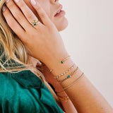 woman in a green blazer with her hand resting against her face and wearing a Zoë Chicco 14k Gold Medium Curb Chain Emerald Cut Emerald Bezel Bracelet layered with three other bracelets and two rings on her hand