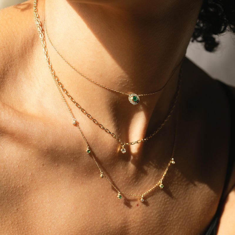 close up of woman wearing Zoë Chicco 14kt Gold Round Emerald and Diamond Halo Necklace around her neck layered with a Floating Diamond Small Square Oval Chain Necklace and a Dangling Emerald and Diamond Necklace with sunlight shining down from the left
