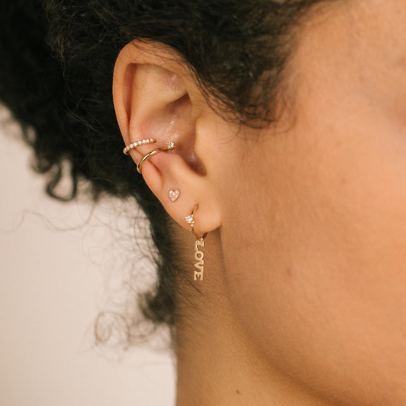close up of woman's ear wearing Zoe Chicco 14k Gold Thick Pave Diamond Ear Cuff and Prong Diamond Thick Ear Cuff layered with an Itty Bitty Pave Diamond Heart Stud and an Itty Bitty Love Charm with Prong Diamond Huggie Hoop Earring