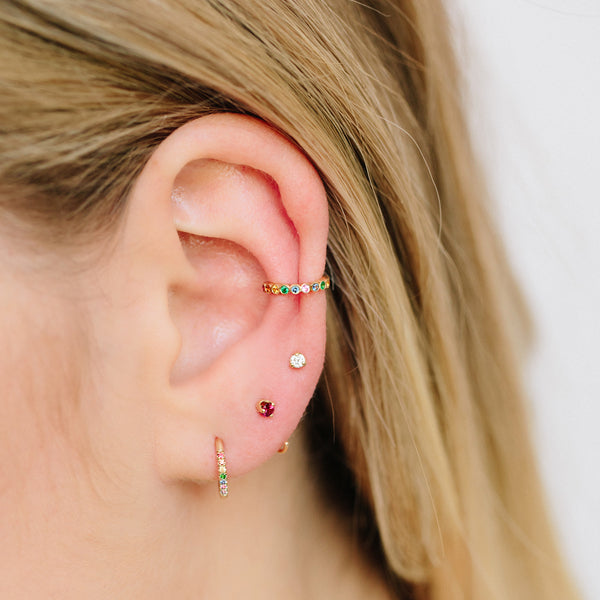 woman's ear wearing Zoe Chicco 14kt Gold 7 Rainbow Sapphires Thick Huggie Hoop Earring with a tiny ruby backwards hoop, a diamond stud, and a rainbow sapphire ear cuff