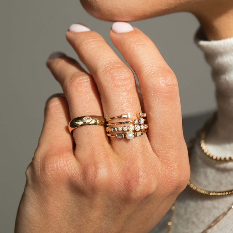 woman holding her hand up against a gray background wearing a Zoe Chicco 14k Gold Half Round Ring with Pear Diamond on her ring finger and a Small Tapered Baguette Diamond Open Ring, 2 Prong Diamond Ring, Graduated Diamond Bezel Eternity Band, and Pearl & Tapered Baguette Ring stacked on her middle finger