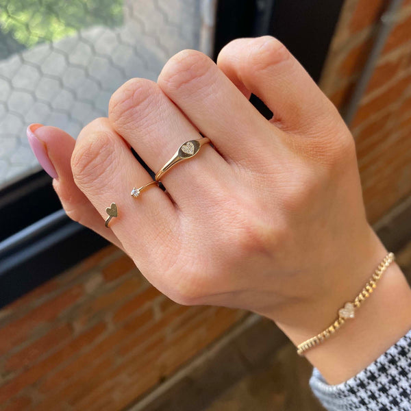 woman's hand against a brick background wearing a Zoë Chicco 14k Gold Itty Bitty Heart & Prong Diamond Open Ring and Pavé Diamond Heart Oval Signet Ring