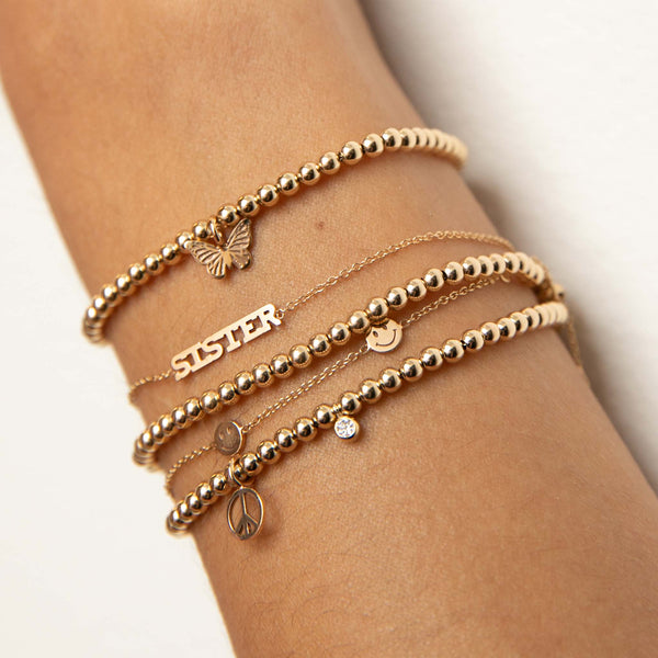 close up of woman's wrist wearing Zoë Chicco 14kt Small Gold Bead Bracelet with Midi Bitty Butterfly Charm layered with an itty Bitty Sister Bracelet, Small Gold Bead Bracelet, 5 Smiley Faces Bracelet, and Small Gold Bead Bracelet with Midi Bitty Peace Sign and Diamond Charms