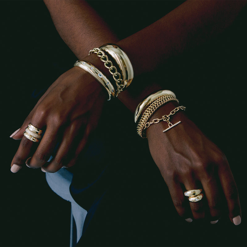 woman's arms crossed on her lap on a dark background wearing Zoe Chicco 14kt gold bracelets and rings stacked together