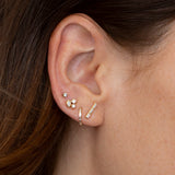 close up of woman's ear wearing Zoë Chicco 14k Gold Channel Set Baguette Diamond Bar Earrings layered with a Baguette Diamond Hinge Huggie Hoop, a single diamond stud, and a mixed cluster stud