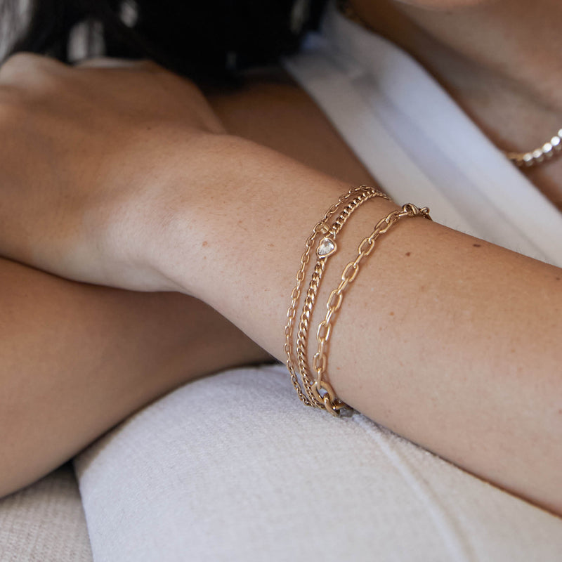 Woman's wrist wearing a Zoë Chicco 14k Gold Floating Heart Shaped Diamond Small Curb Chain Bracelet layered with two heavy chain bracelets