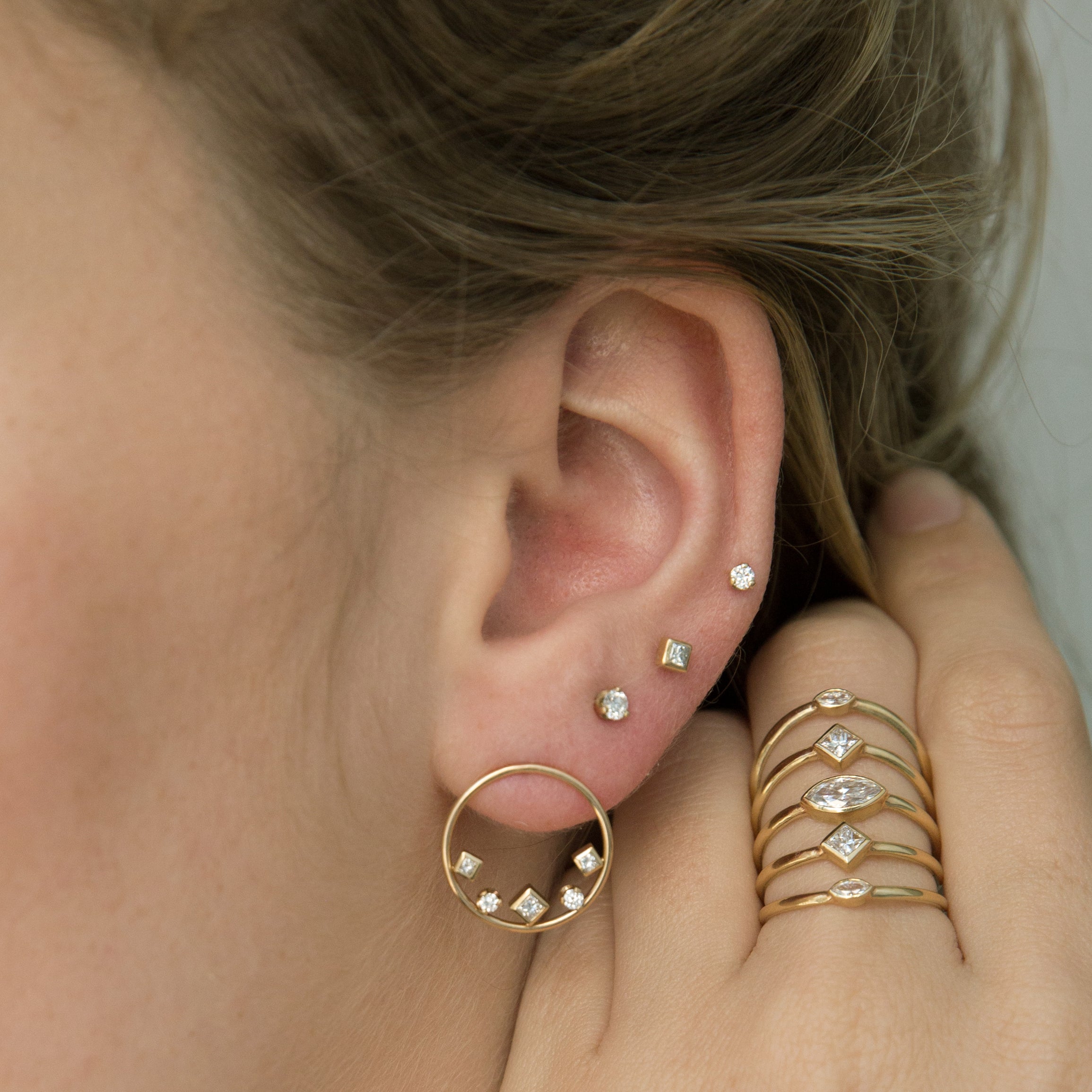 Close up of a woman's ear and hand wearing Zoe Chicco 14k gold and diamond rings and earrings on a neutral background