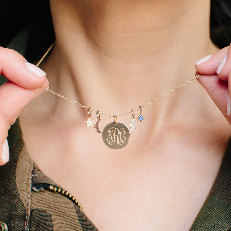 woman holding up a chain with Zoë Chicco 14kt Gold Midi Bitty Moon Charm Pendant, a monogram pendant, a blue sapphire bezel charm, pave diamond star charm, and a pave diamond lightning bolt charm