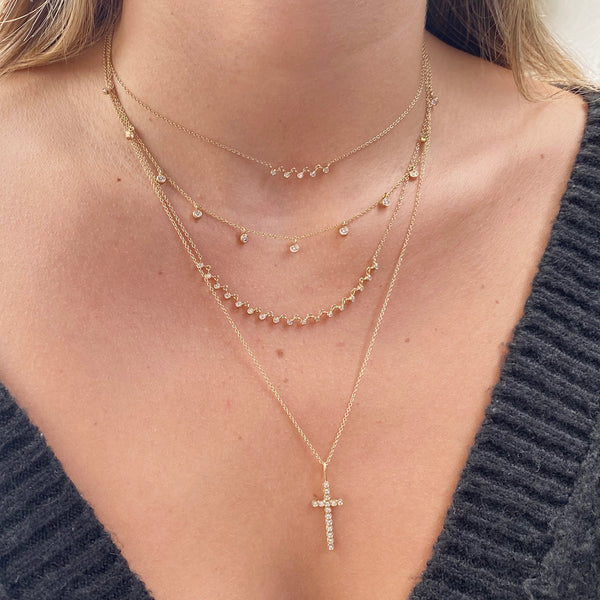 woman in sweater wearing Zoë Chicco 14kt Gold Linked 5 Diamond Bezel Necklace layered with a Diamond Cross Pendant Necklace, Floating Diamond Station Necklace, Linked 17 Diamond Bezel Necklace, and a Long Diamond Cross Pendant Necklace
