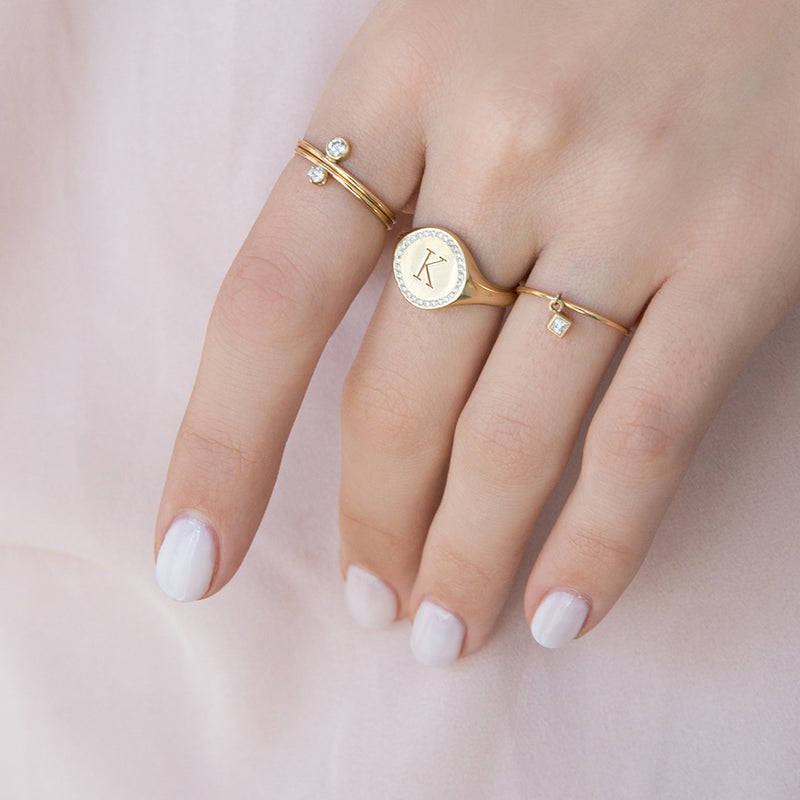 woman's hand on light pink background wearing Zoë Chicco 14kt Gold Round Initial Signet Ring with Diamond Halo, a Princess Diamond Charm Ring, and two single diamond band rings